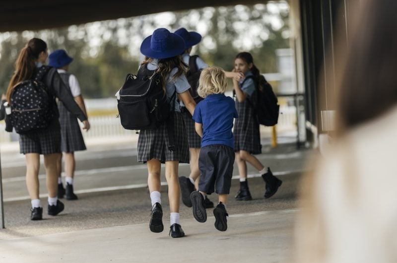 A schoolgirl holds a younger boy's hand as they walk toward other children outside school.